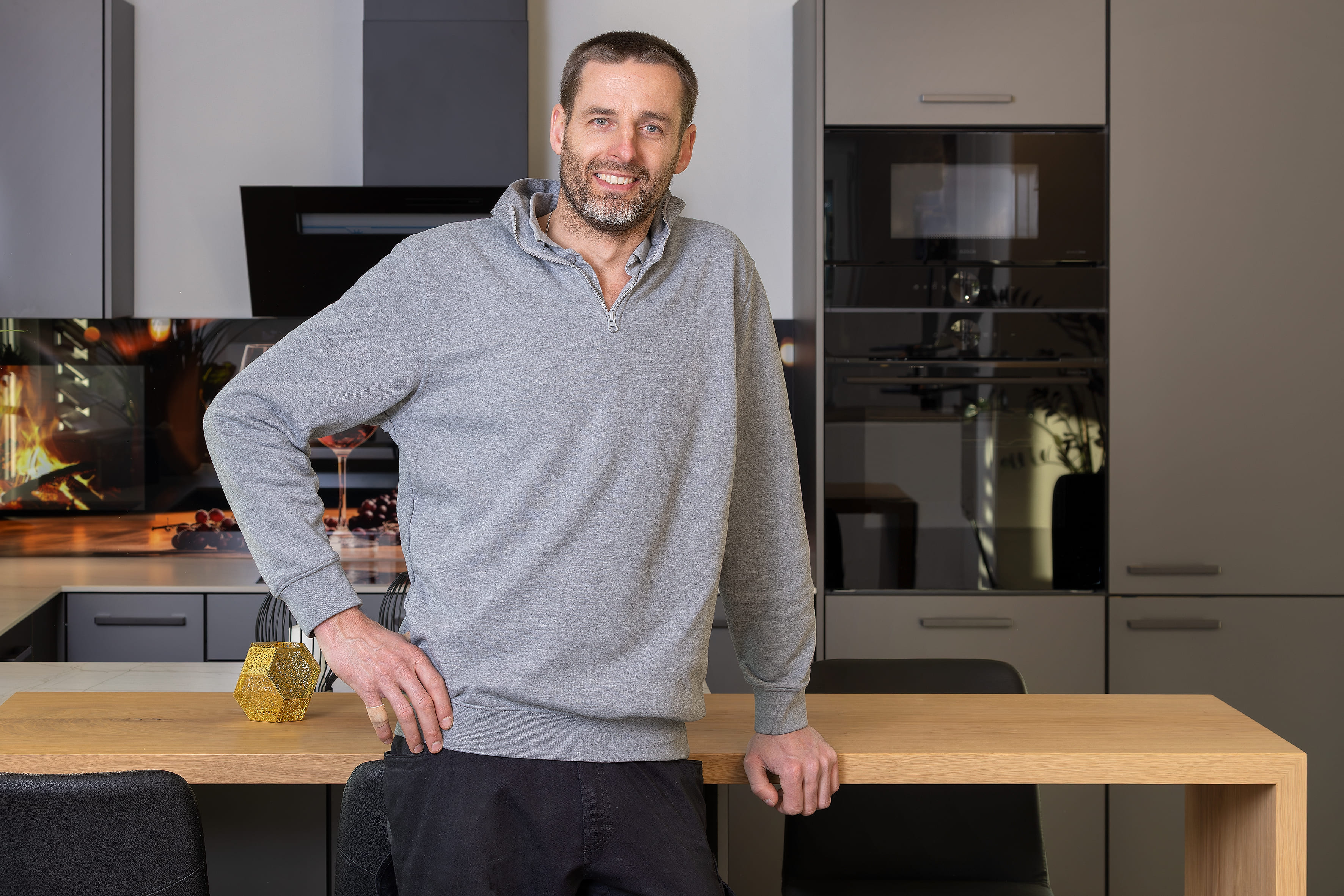 Mann mit grauem Pullover lächelt in moderner Küche, lehnt an einem Holztisch.