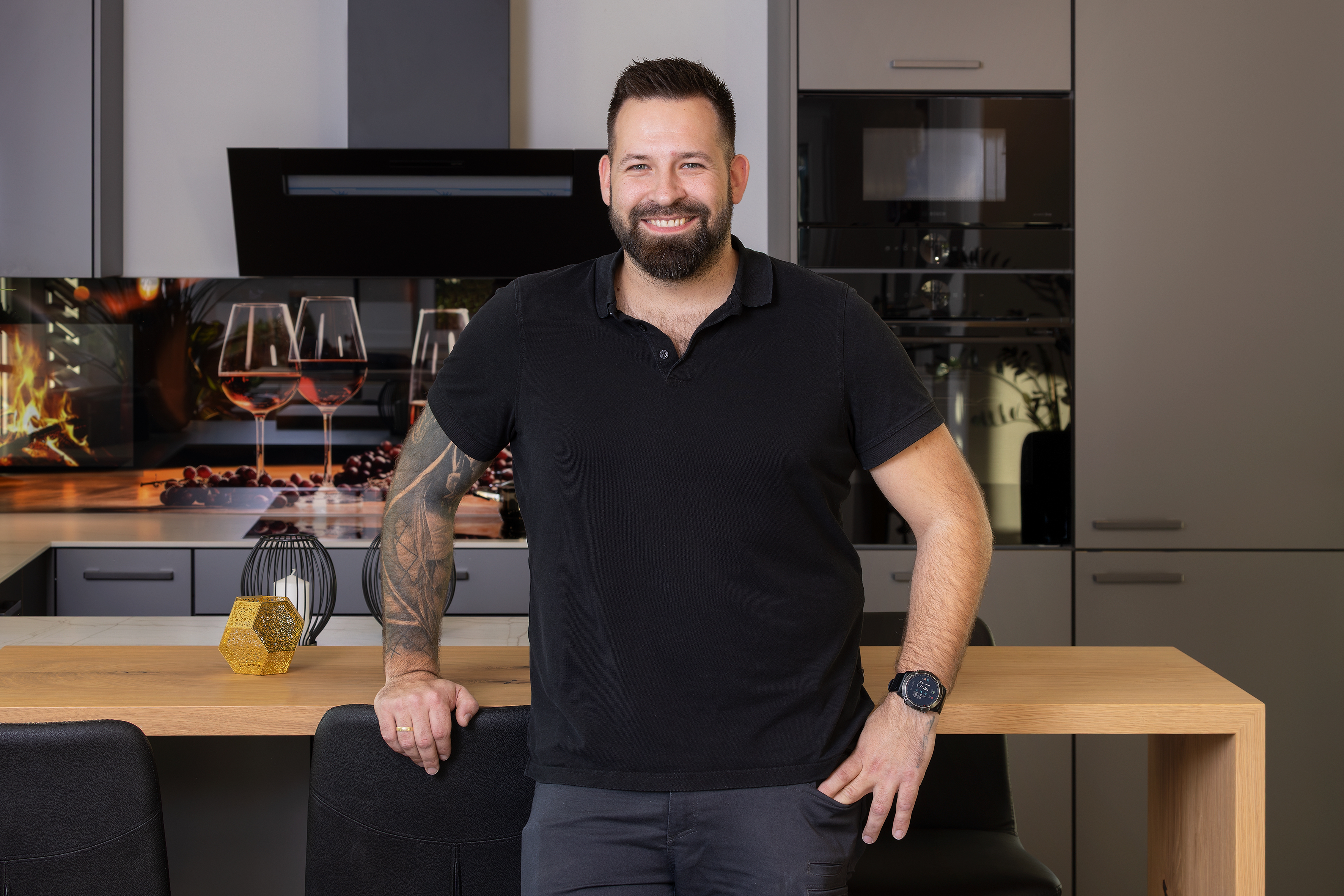 Mann mit Bart und Tätowierung lächelt in moderner Küche, lehnt an Esstisch.