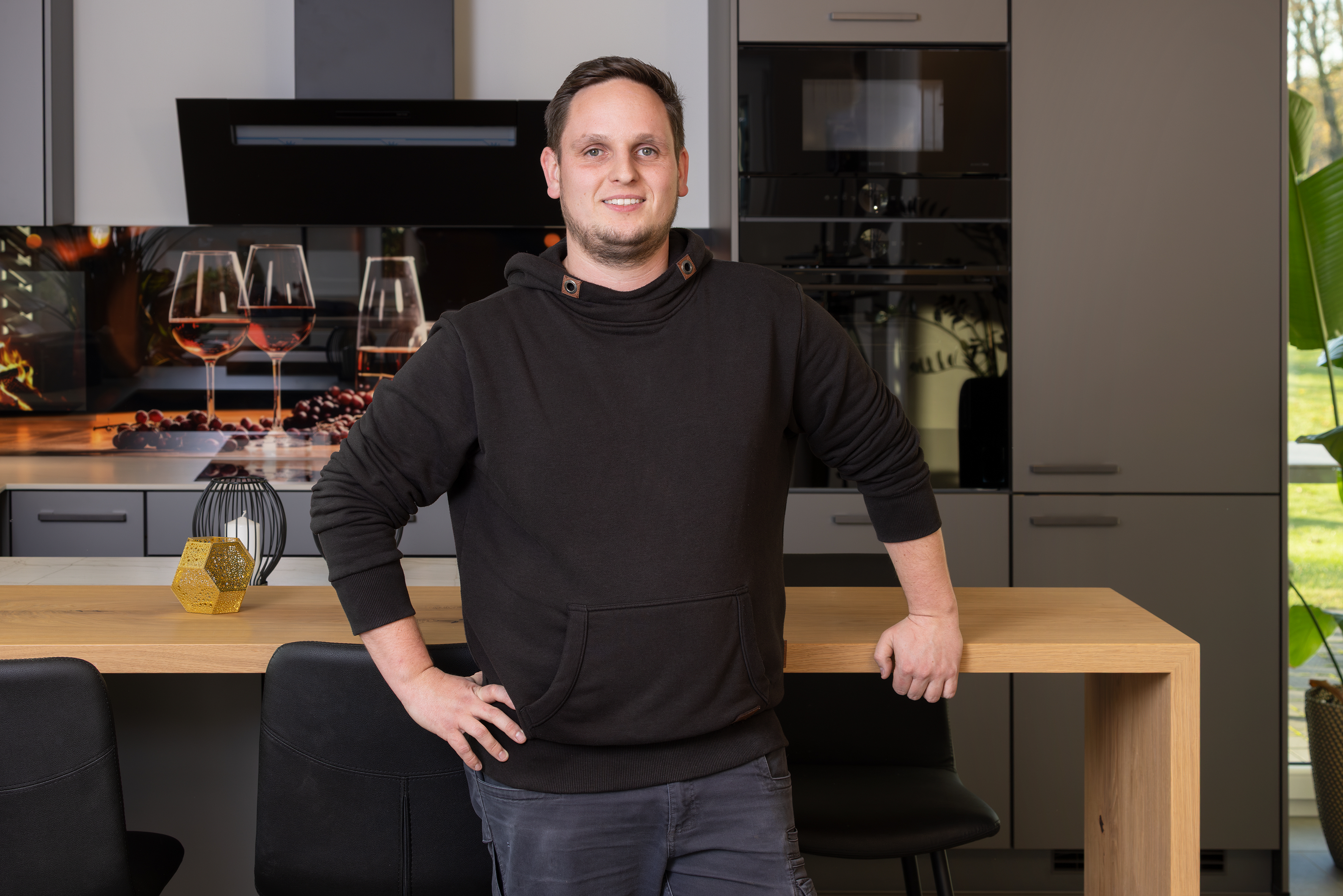 Mann steht lächelnd an einem Holztisch in einer modernen Küche.