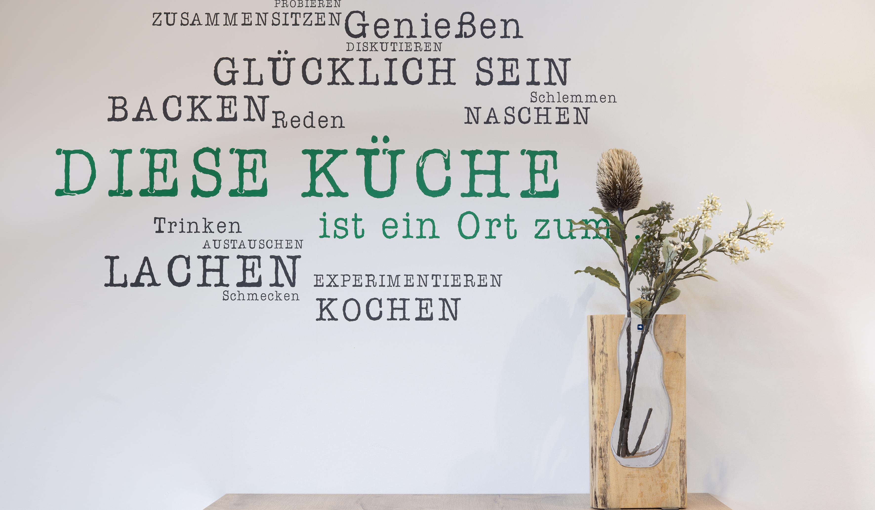 Küchenwand mit Sprüchen und Vase mit Trockenblumen auf einem Holztisch.