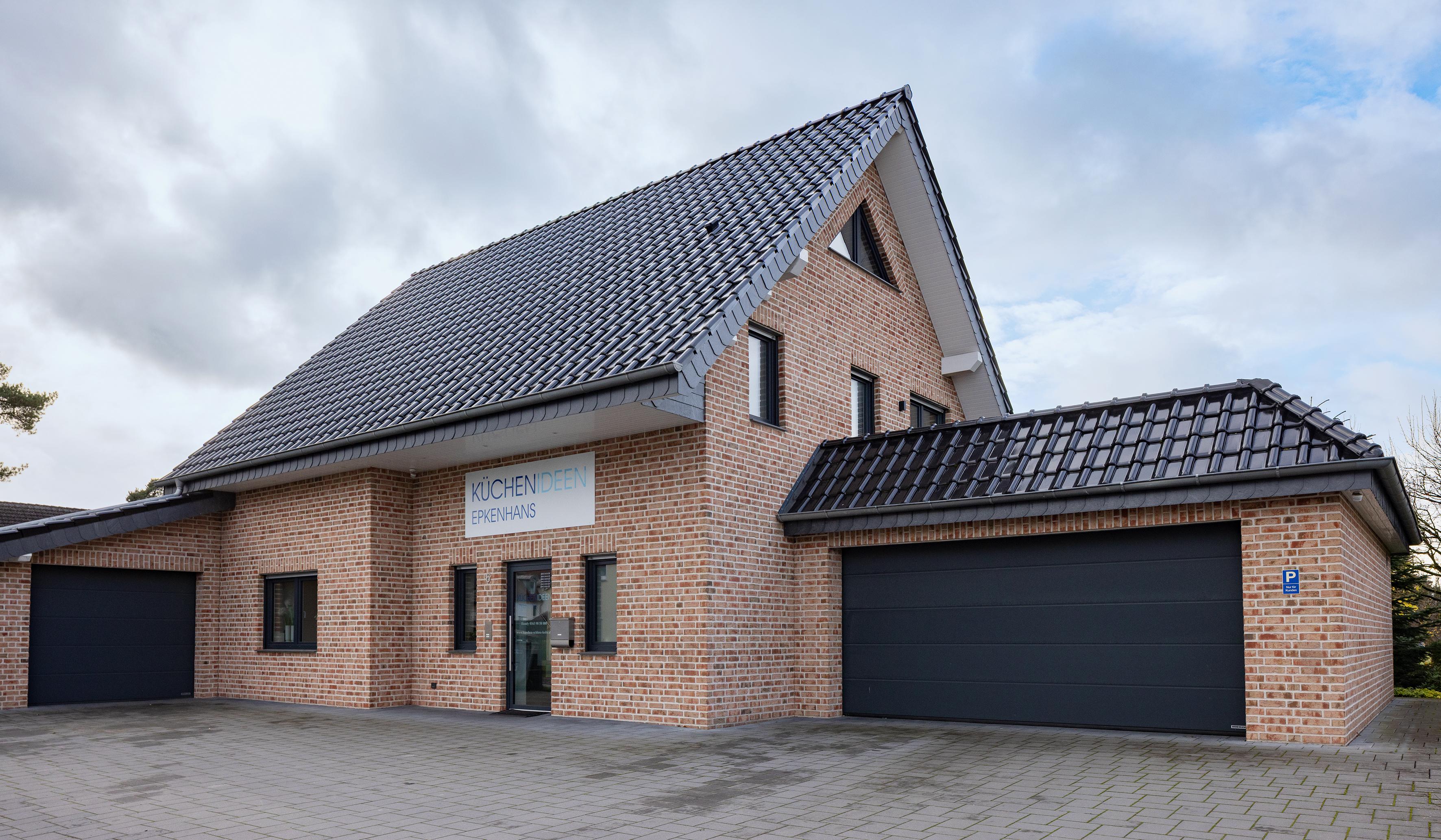 Zweistöckiges Backsteingebäude mit dunklem Dach und zwei großen Garagentoren. Küchenstudio Epkenhans Außenansicht. 