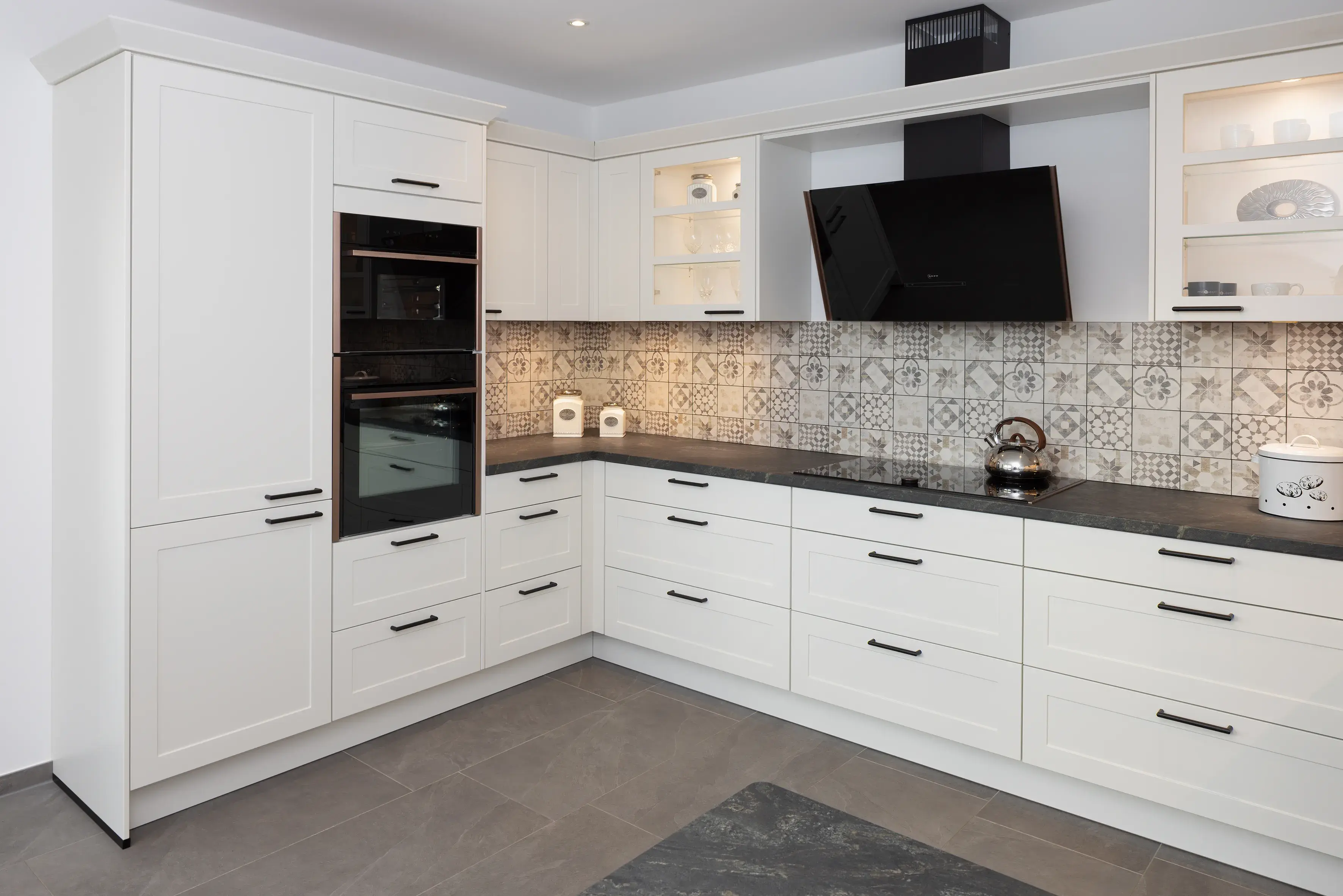 Moderne, weiße Einbauküche mit gemustertem Fliesenspiegel und schwarzen Elektrogeräten.