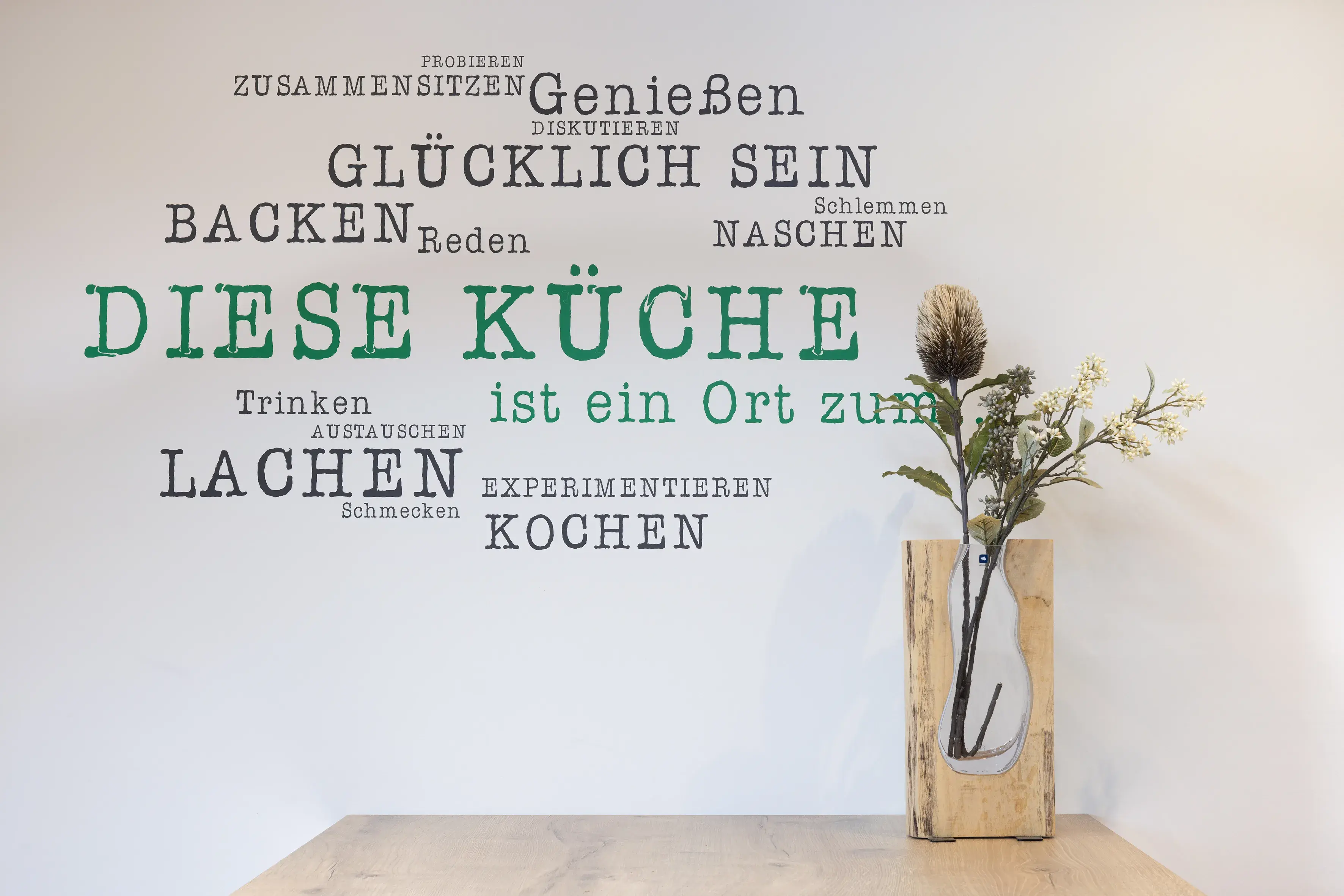 Küchenwand mit Sprüchen und Vase mit Trockenblumen auf einem Holztisch.